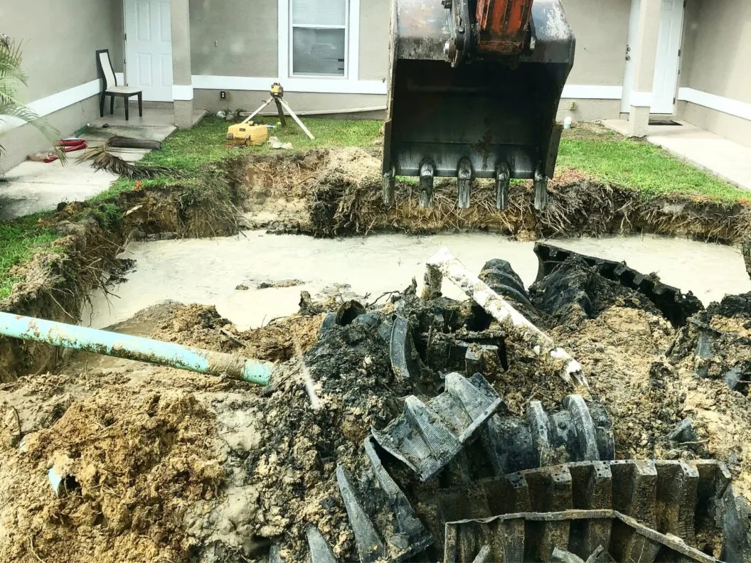 Excavator bucket over a flooded pit where crushed old drain field chambers and a pipe were dug out beside a house, drain field repair