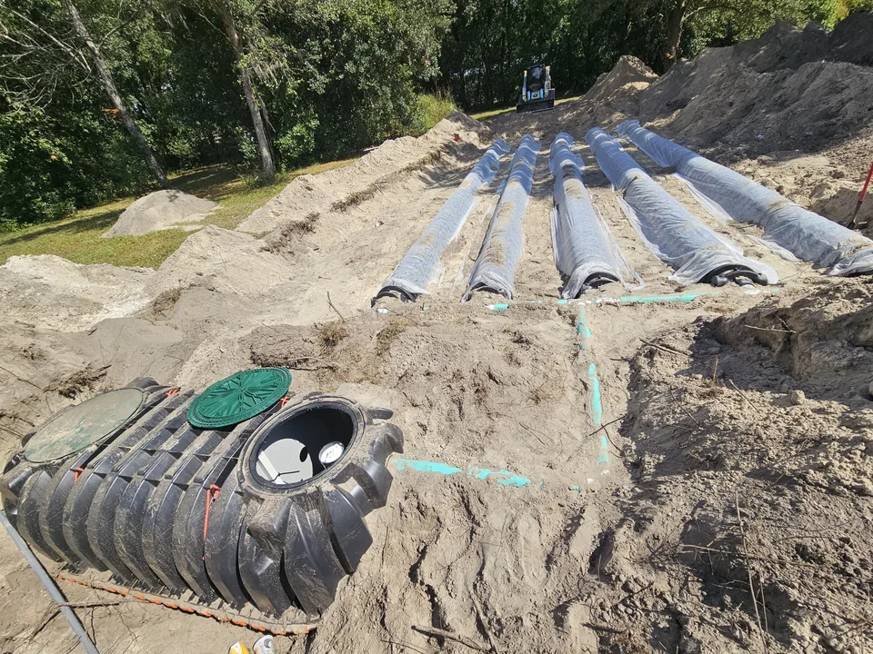 Drain field lines under plastic sheeting in a sandy dig beside a black septic tank, drain field repair
