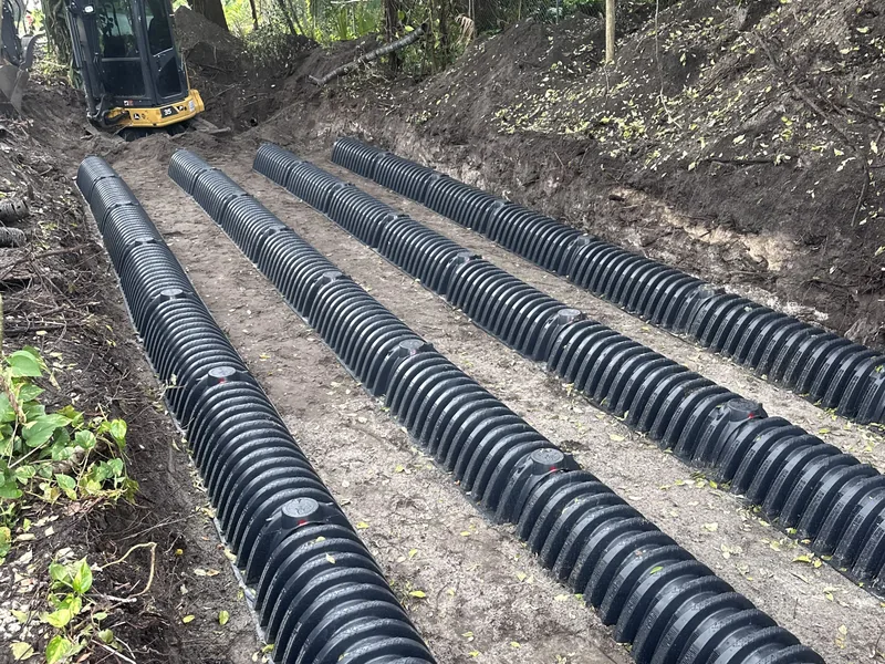 Drain field chambers in an open trench under trees with a mini excavator behind, emergency septic service