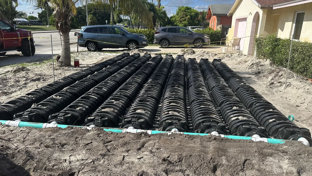 Rows of black drain field chambers with a green distribution pipe in a sandy front yard beside a house, septic system installation