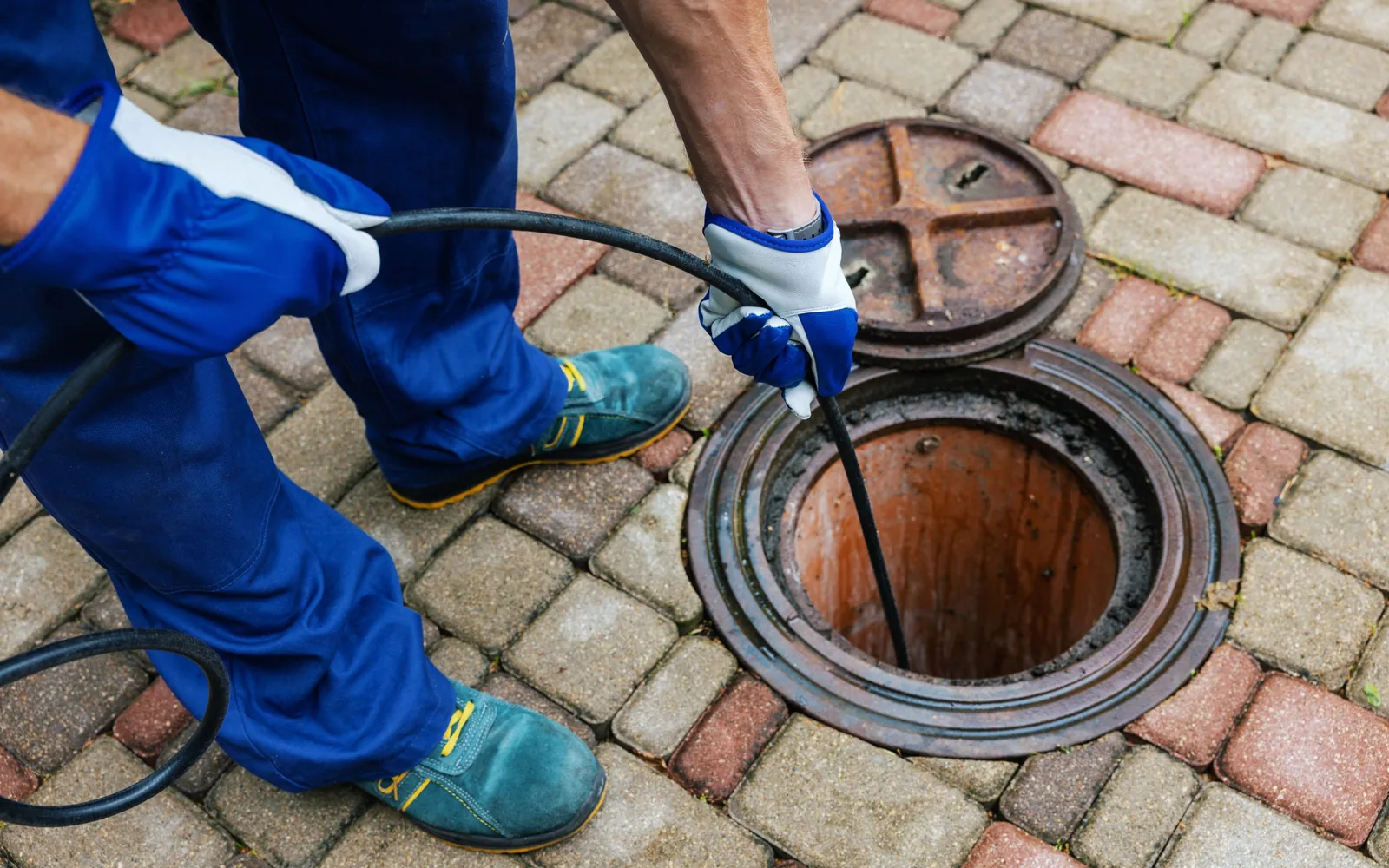 Gloved hands feeding a black jetter hose down an open sewer access in a brick paver drive, hydro jetting