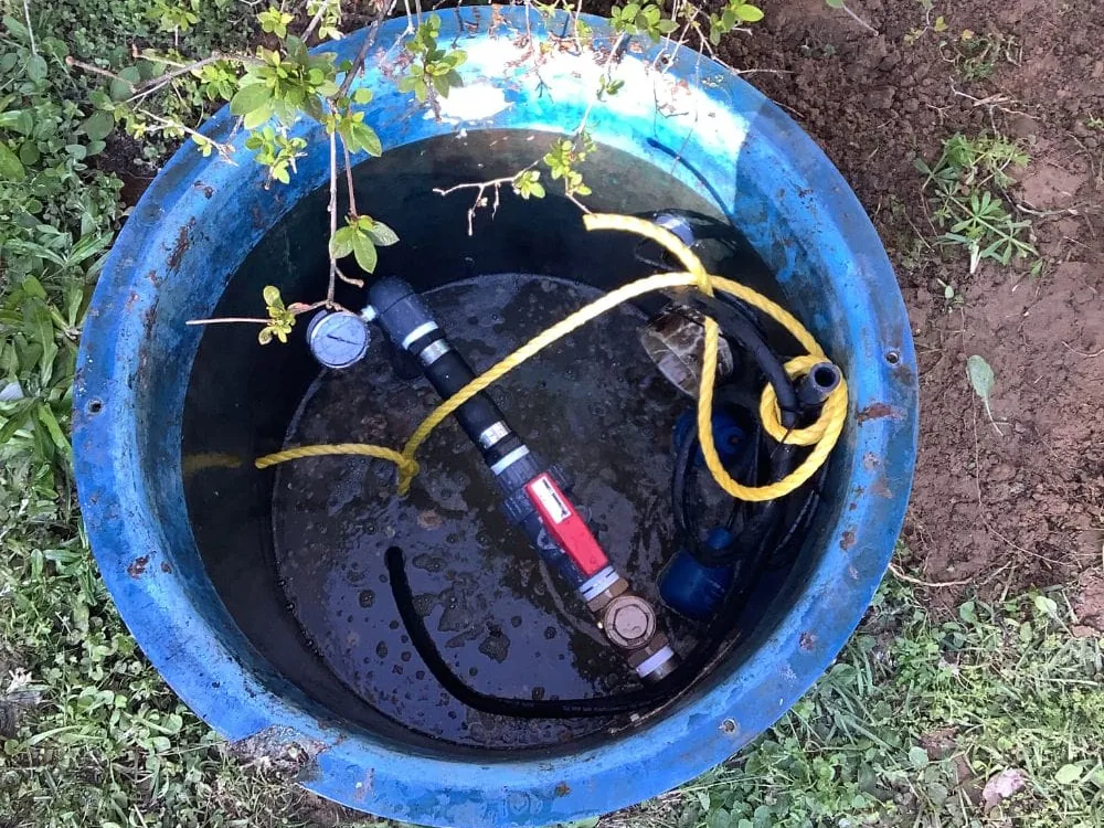 Open pump basin in a yard seen from above with the submersible pump, discharge pipe, valve and gauge inside, lift station pump service