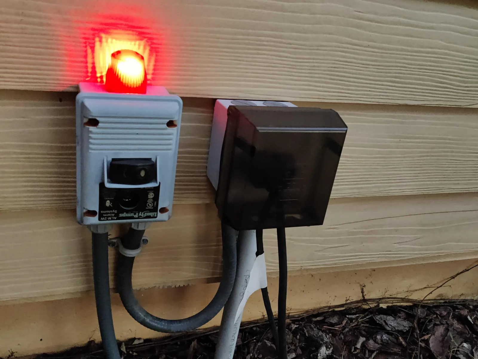 Pump high water alarm with its red beacon lit, mounted on a house wall beside an outdoor junction box, lift station pumps