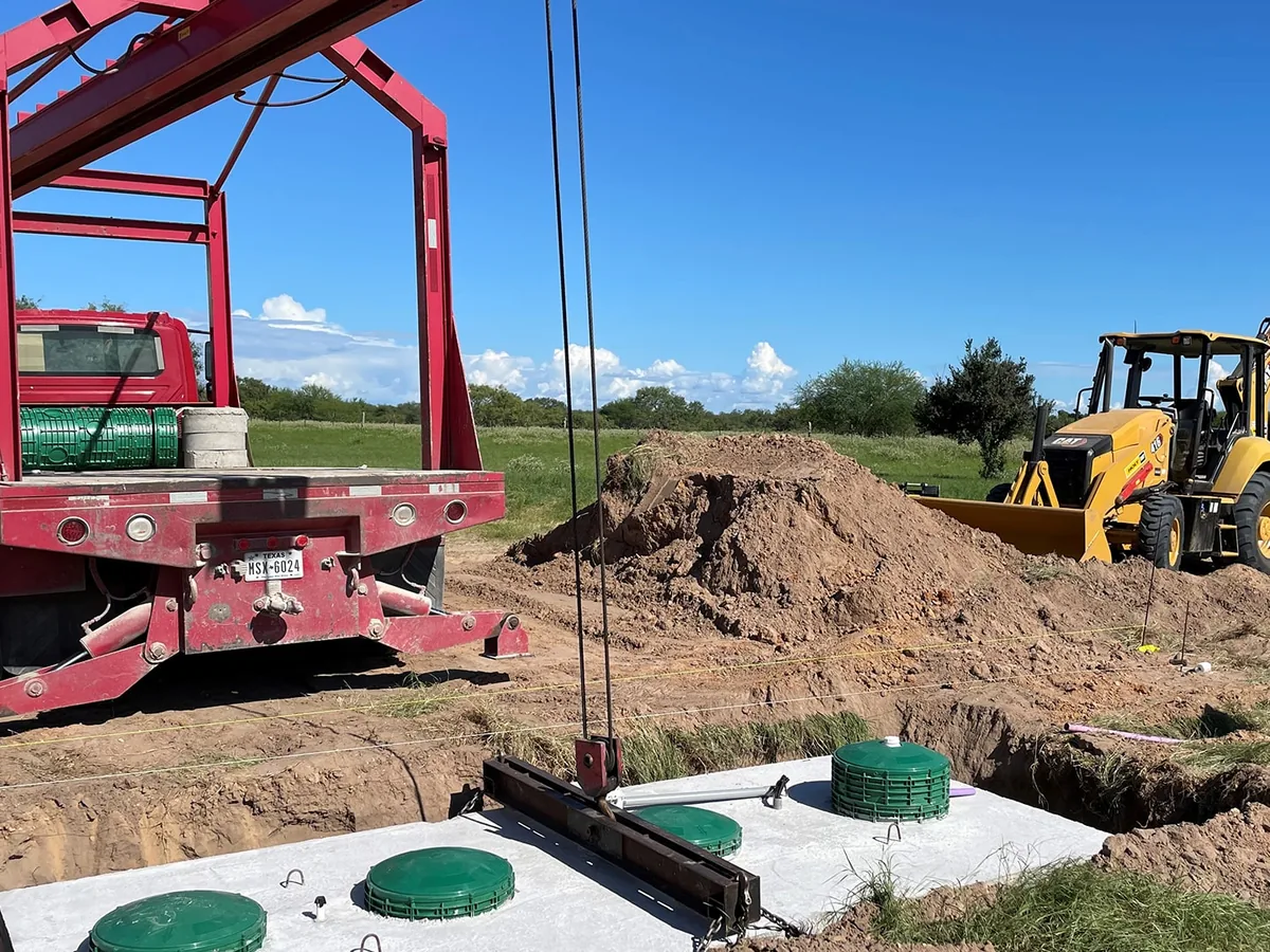 Boom truck setting a concrete septic tank with green risers into a dug hole beside a backhoe, septic system installation