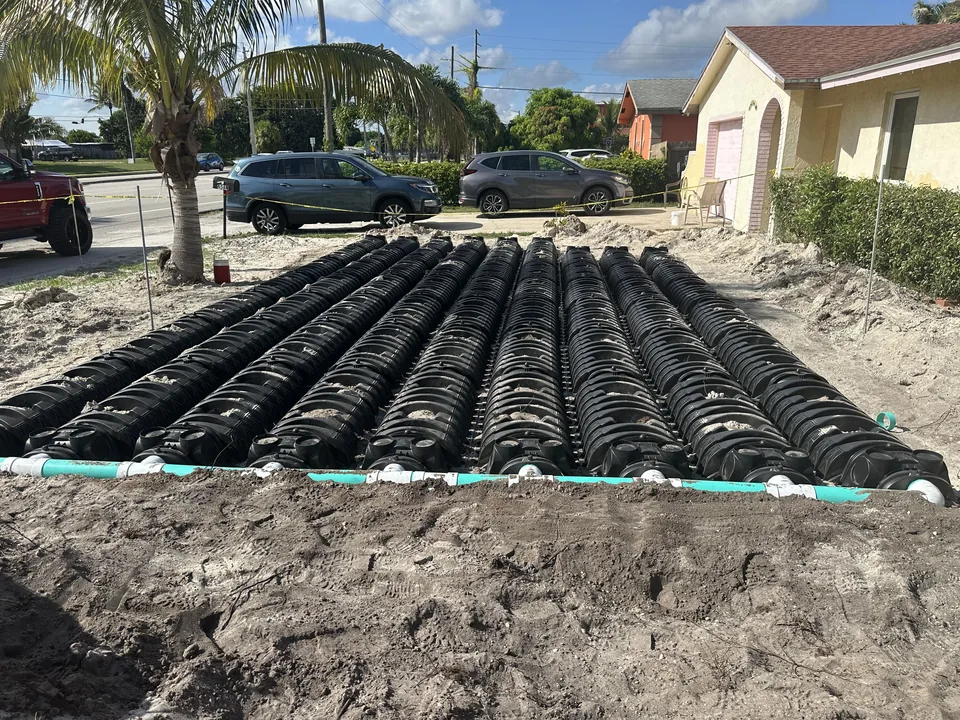 Rows of black drain field chambers joined by a green distribution pipe in a sandy front yard, septic system installation