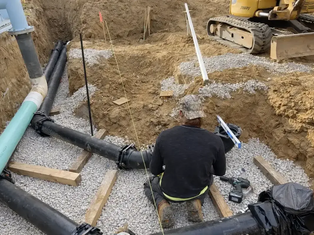 Worker joining black drain pipes on a gravel bed in an open trench, septic system installation