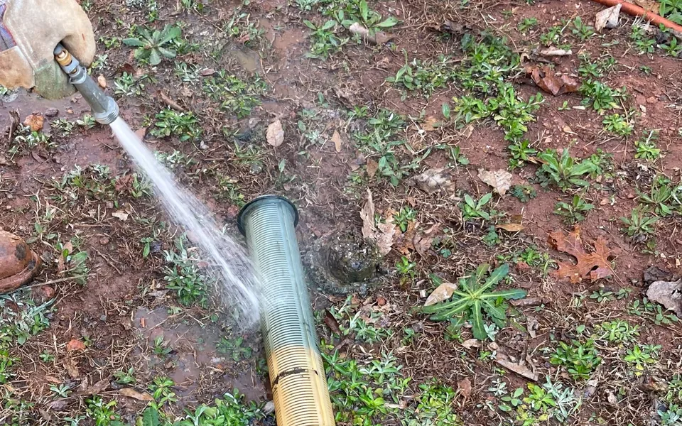 Gloved hand hosing sludge off an effluent filter cartridge on the ground beside the tank opening, septic tank filter cleaning