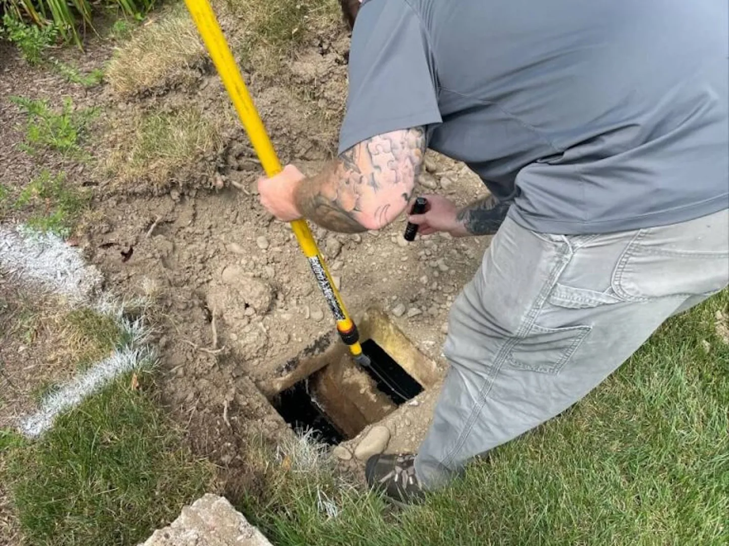 Technician pushing a probe pole into an open concrete septic tank access with a flashlight in his other hand, septic tank inspection