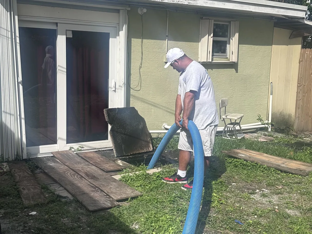 Concrete septic tank lid set against a house wall while a technician feeds a vacuum hose into the tank, septic tank lids and risers