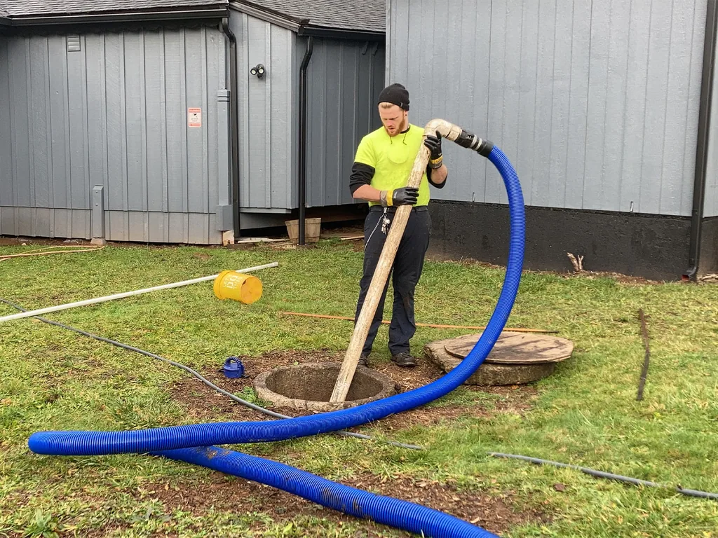 Technician lowering a vacuum hose into an open septic tank on a lawn, septic tank pumping