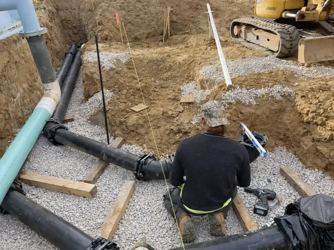 Worker joining black pipes on a gravel bed in a dug trench beside a mini excavator, septic tank repair